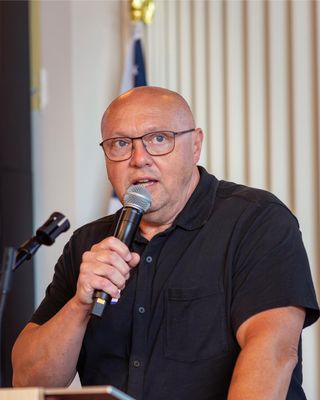 A bald man with glasses holding a microphone and speaking at a podium, he looks focused and there's an American flag just visible behind him.